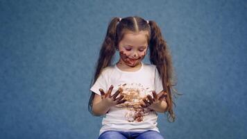 Happy kid with dirty hands and face by chocolate. Funny little girl has smeared face, hands and white T-shirt with chocolate in the studio. video
