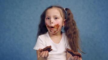 Dirty face of a girl eating chocolate. Two pieces of sweet chocolate in child's hands. Portrait of a little kid with smeared face isolated in studio. video