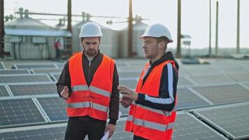Technicians on photovoltaic farm of a renewable energy. Two workers in safety helmets are talking on the background of solar panels. video