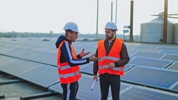 Installing photovoltaic panels. Workers in orange uniform and hardhats on solar farm background. Engineers are talking among solar panels. video