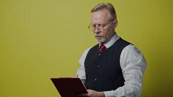 Portrait of serious grey-haired man with a folder. Senior man in glasses in suit writing on a folder. Old man expresses different facial emotions on yellow background. video