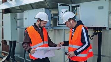 Engineers discuss the further work with a paper plan. Male workers in safety helmets looking at the plan scheme on the electrical equipment background. video