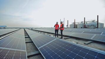 Engineers talk on the solar farm background. Rows of solar panels and two workers in orange uniform and hard hat. Installation of innovative sunny batteries. video
