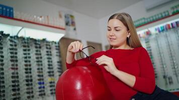Young woman buyer choosing medical glasses. Attractive girl sitting in the optical store and looking at a pair of trendy eyeglasses. Eyewear choice. video