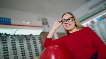 Beautiful woman in stylish eyeglasses. Female customer sitting in armchair and trying on new glasses in optical store. Eyewear concept. video