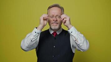 Funny grey-haired man putting on glasses. Senior man in elegant clothing with eyeglasses making amusing eyes on yellow background. Slow motion. video