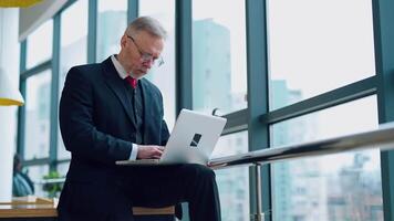Senior businessman in suit working near office window. Elderly elegant man is typing on a laptop while sitting near the window. Business concept. video