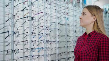 Beautiful girl choosing new spectacles. Smiling blonde woman trying on stylish eyeglasses in the optics store. Eyewear concept. video