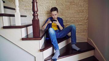 Teen boy playing on the phone on stairs. Boy sitting on a stairs at home and playing games on his mobile phone. Gaming concept. video
