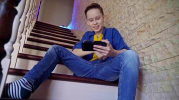 View from below on a boy sitting on stairs at home. Happy teenager playing on his mobile phone and wins the game. Gaming concept. video