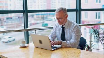 Senior freelancer using laptop working from home. Smiling grey-haired man in glasses typing on computer and drinks coffee enjoying distant job. video