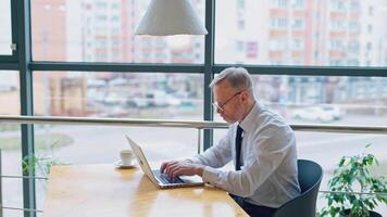 Side view of a senior man typing on a laptop. Successful businessman in white shirt and tie sitting at the desk and working near the window. video