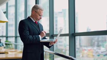 Mature senior serious businessman with laptop in hands thinking while checking e-mails standing near window with panoramic city view. from the side. video
