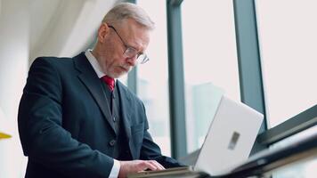 Senior serious businessman standing near window with panoramic city view. Laptop in hands, man thinking while checking e-mails standing near window. from the side. video