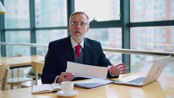 Businessman is checking a printed report or document while sitting at table, working on paper work at a laptop explaining his point of view. in an office. video