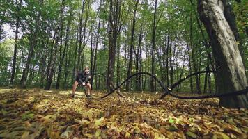 Sportive man exercises with battle ropes in forest. Sportsman doing waves with cables while training in the autumn daytime. Slow motion. video