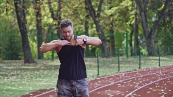 Young man takes off his jersey at the open stadium. Muscular sportsman shows his pumped body against the background of green trees. video