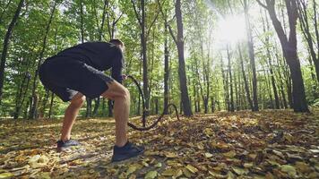 Strong man doing waves with ropes during workout. Backside view of a young athlete exercising with black ropes among trees in the forest. Slow motion. video