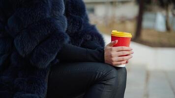 Female hands hold a cup of beverage outdoors. Beautiful woman in a fur coat sitting in the park and drinking from plastic glass. Slow motion. video