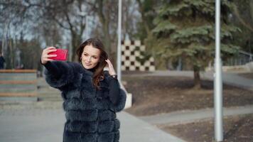 Portrait of a beautiful girl doing selfie in the park. Young woman in luxury fur coat doing photo of herself on the red phone in a sunny winter day. video
