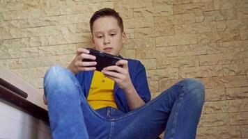 Portrait of a boy with a smartphone at home. Teenager is playing on his phone while sitting on stairs. Social distancing and self isolation in quarantine. video