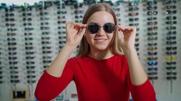 Portrait of smiling girl in black sunglasses. Attractive young woman is trying on stylish eyeglasses on the background of new spectacles at optical store. video