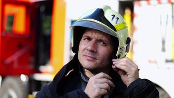 Young fireguard in protective uniform putting helmet against background of a big red car. Portrait of young confident fireguard in protective uniform against background of fire truck. Slow motion video