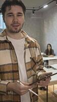 Handsome university student with dark brown hair is writing on electronic tablet during break from lectures in middle of classroom. Vertical. video