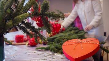 Red Christmas tree toy in shape of heart hangs from branch, with box of chocolate next to it. These are sweet Valentine's Day chocolates. In background, decorators create winter scene with fir branches. video