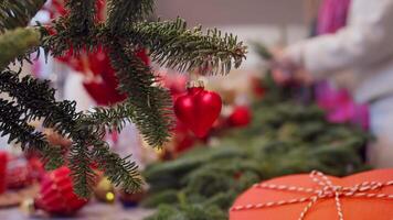 Heart-shaped toy, symbol of love, is hanging from branch. Behind it, florists are creating decorations for homes using fir branches during holidays such as Valentine's Day and Christmas video