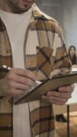 A close-up of the hands of a young man, a student wearing a plaid shirt, in a classroom, using a tablet and stylus for studying. Vertical. video