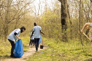 Diverse group of environmentalists collecting rubbish in trash bags, picking in junk and plastic waste from the forest. Activists cleaning the woods from garbage, nature pollution. photo