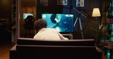 Player in cozy living room enjoys game with spaceship shooting laser streams using crosshair overlay to hit enemies. Man plays on console system with Ray tracing graphics and high FPS, zoom out shot photo
