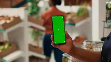 Vendor showing phone with greenscreen layout at supermarket, holding blank copyspace mockup on display. African american seller working at local grocery store, isolated chromakey screen. photo