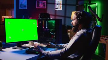 Man in dimly lit apartment playing games on isolated screen gaming PC at computer desk, enjoying day off from work. Gamer battling enemies in online multiplayer shooter on chroma key monitor photo