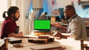 BIPOC artisan and apprentice compare wood art piece with schematic displayed on green screen laptop. Woodworking experts in studio crosschecking wooden object with mockup notebook blueprint, camera A photo