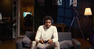 African american man playing shooter videogame in cozy living room, holding controller. Gamer participating in PvP online multiplayer game using console system, relaxing at home photo