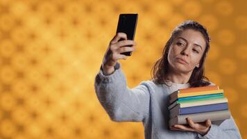 Smiling woman holding pile of books, enjoying reading hobby, taking selfies with cellphone. Cheerful lady with stack of novels in arms doing pictures with mobile phone, studio background, camera A photo