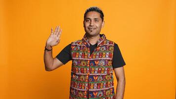 Portrait of jolly man smiling and waving hand, saluting, gesturing, isolated over studio backdrop. Joyous upbeat indian person in traditional attire doing greeting hand gesture, camera B photo