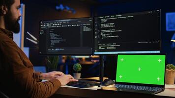 Programmer coding on computer next to isolated screen laptop in home office, developing software application. Teleworking IT specialist doing programming on PC near chroma key notebook, camera B photo