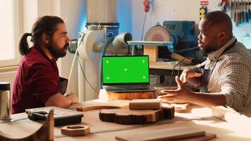 Woodworker looking over blueprints on green screen laptop in CAD software with apprentice. Craftsperson and coworker designing custom 3D furniture on chroma key notebook in studio, camera A photo