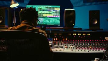 African american audio engineer using sound mixing console for managing tracks, music post production. Producer pressing motorized control knobs, looks at editing software. Camera B. photo