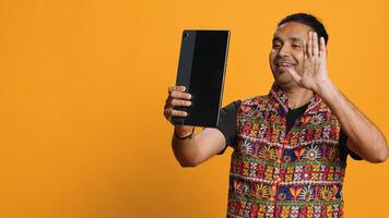 Radiant man saluting friend over online videocall, doing friendly gestures. Joyful person communicating with mate on teleconference using tablet, studio background, camera A photo