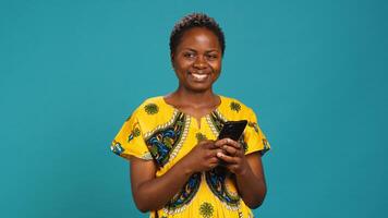 Native girl in ethnic outfit browsing internet webpages in studio, using her smartphone to check texts and social media platforms. Young person standing against blue background. Camera A. photo