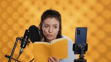 Influencer filming reading book with smartphone on tripod, creating audiobook, studio background. Woman vlogging with mobile phone while producing digital recording on novel using microphone, camera A photo
