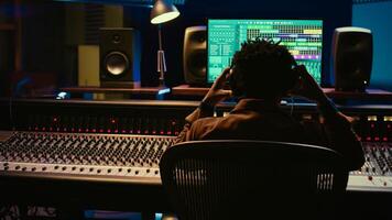 African american sound engineer recording and editing tracks in post production, using mixing console with knobs and switchers. Skilled audio technician producing new music in studio. Camera B. photo