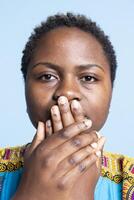 African american woman covering her mouth in front of the camera, being silent and recreating three wise monkeys symbol. Female model showing motivational sign and wisdom in studio. photo