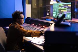 African american record producer composing tunes in control room, editing music and adjusting volume levels with panel board buttons. Audio expert pressing sliders to add sound effects. photo