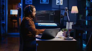 Developer focused on typing complex code in neon lit home office, listening music and developing software application. IT expert wearing headphones, solving coding project tasks on laptop, camera B photo
