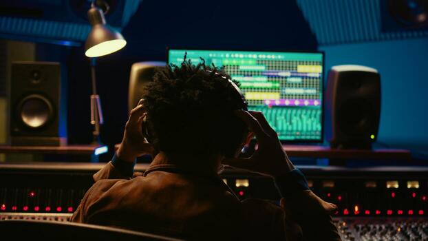 African american music producer recording tracks and editing them in post production, using mixing console with knobs and sliders. Audio expert processing sounds with technical gear. Camera B. photo